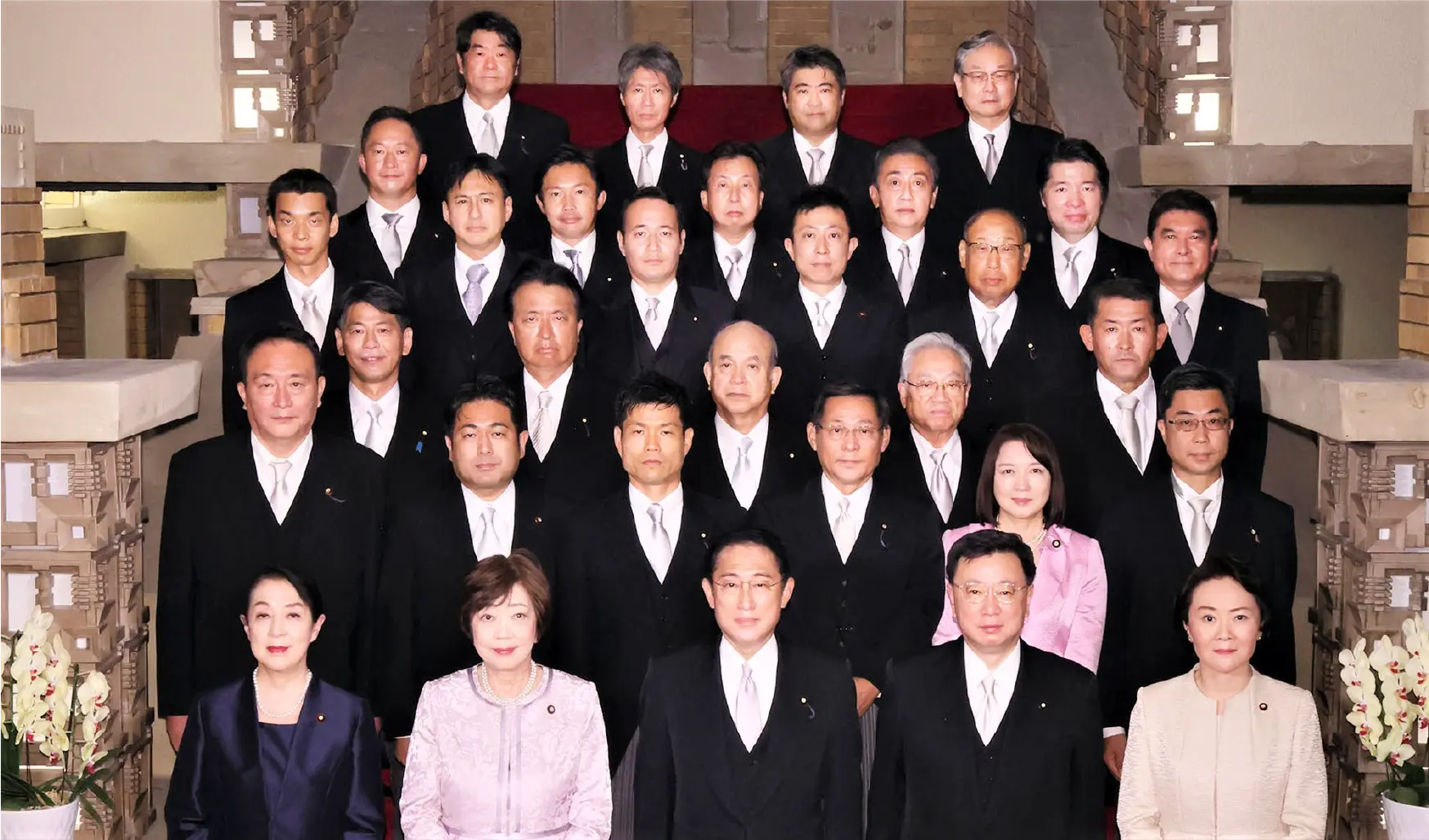 岸田改造内閣の集合写真。正装した閣僚たちが段状に並び、中央最前列に岸田文雄首相が立つ。かつまた孝明は岸田首相の左上に立っている。背景には歴史的建築様式の建物内部が映る。