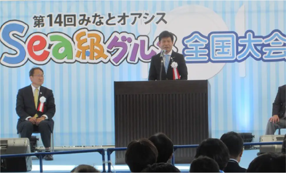 「第14回 Sea 級グルメ全国大会」の看板が背景に立つ壇上でかつまた孝明が挨拶をしている様子。壇上には他の議員や関係者も座っており、会場には多くの人々が集まっている。