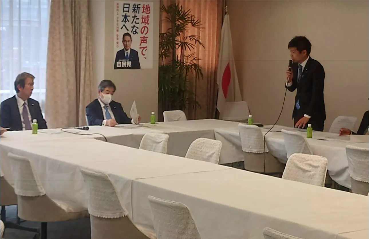 自民党半島振興小委員会にて、かつまた孝明(画像右)が立ち上がりマイクを持って話す様子。画像左には他の議員たちが座り話を聞いている。