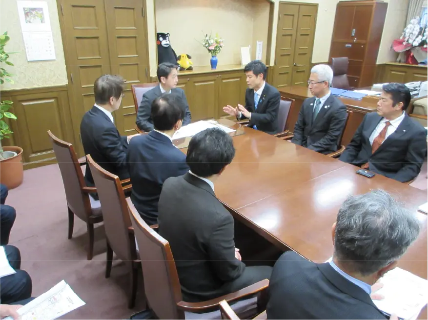 財務省の会議室にてかつまた孝明が要望をする様子。周りには他の関係者たちが座り、真剣な表情で話を聞いている。