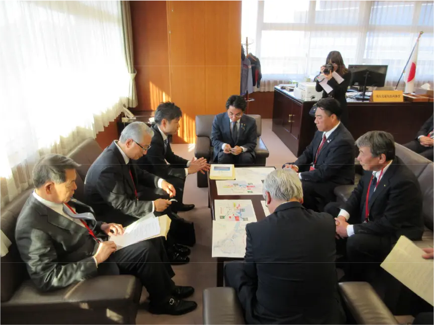 国土交通省の会議室にてかつまた孝明が要望をする様子。周りには他の関係者たちが座り、資料を広げながら真剣な表情で話を聞いている。