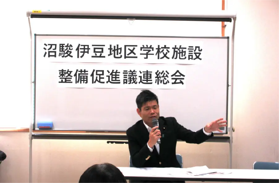 「沼駿伊豆地区学校施設整備推進議連総会」と書かれたホワイトボードの前で、かつまた孝明がマイクを持って話している様子。