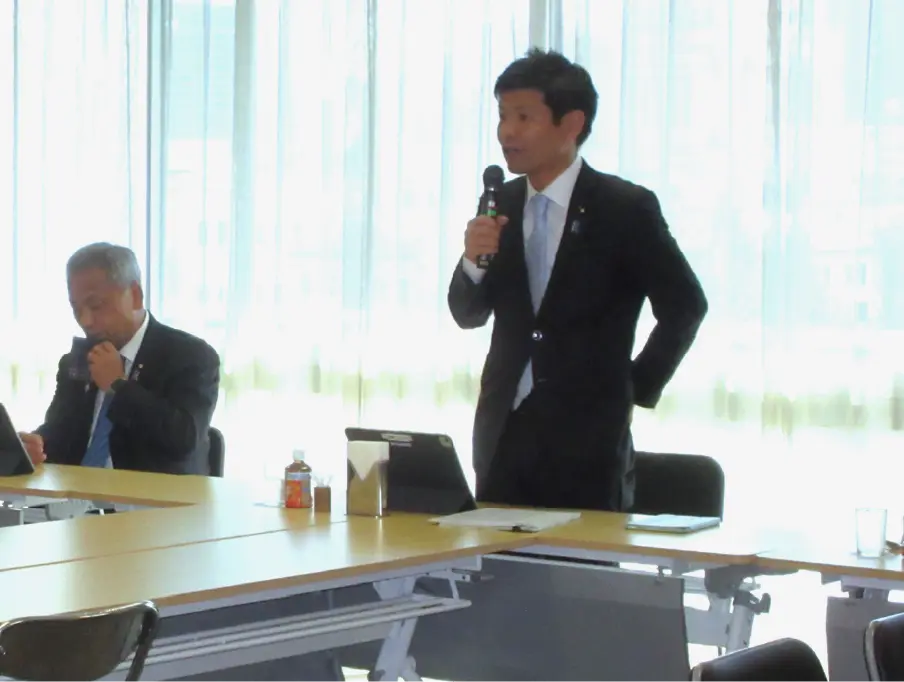 自民党本部にて、部会長のかつまた孝明が立ち上がりマイクを持ち強く訴える様子。