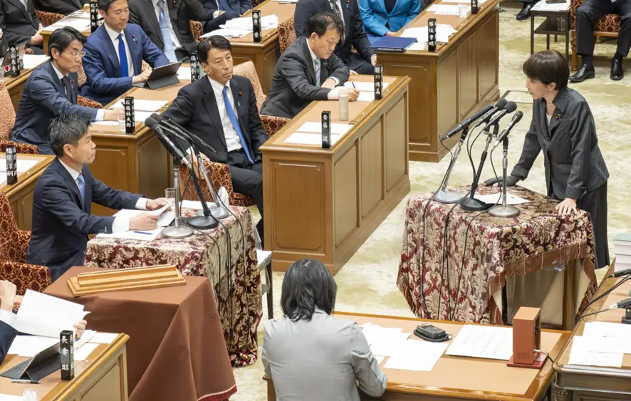 衆議院予算委員会にてかつまた孝明がマイクの前に立ち資料を確認しながら、高市総理に質問をする様子。