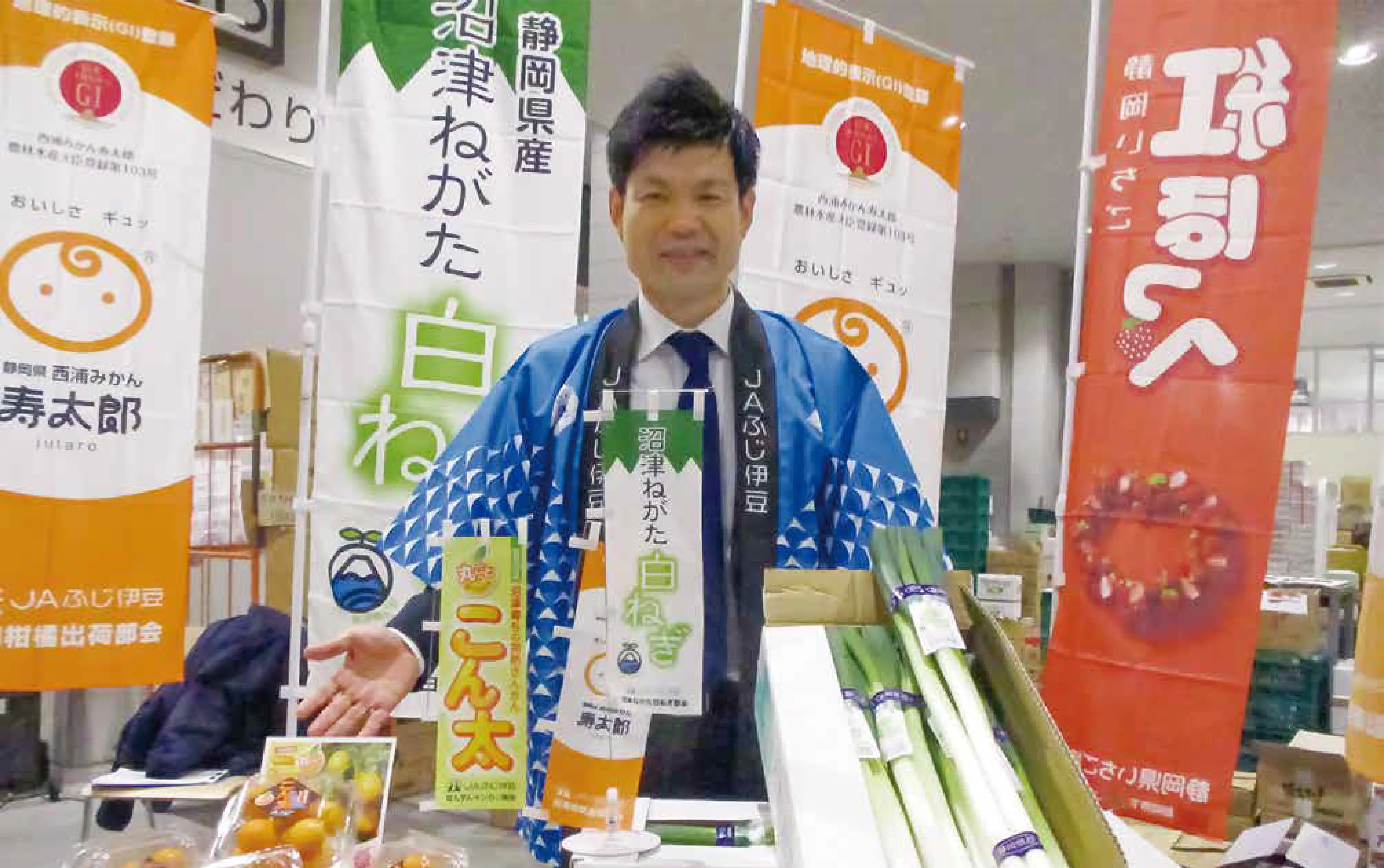 豊洲市場の特設会場にて「JAふじ伊豆」と書かれた法被を羽織り、かつまた孝明が静岡の食の魅力を発信している様子。写真手前には白ネギ・みかんが並び、背面には白ネギ・みかん・イチゴなどの静岡の特産物の旗が置かれている。